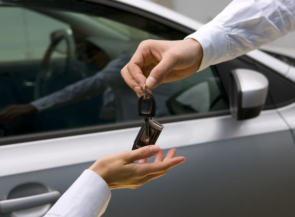 Mann gibt Frau einen Autoschlüssel in die Hand, im Hintergrund ein Auto