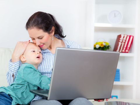 Junge berufstätige Frau mit einem Laptop auf dem Schoß und einem Kind im Arm